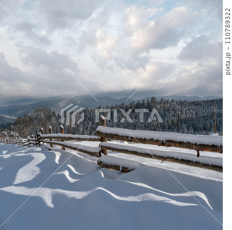 Picturesque waved shadows on snow from wood fence. Alpine mountain winter hamlet outskirts, snowy path, fir forest on far misty and cloudy hills. Picturesque waved shadows on snow from wood fence. Alpine mountain winter hamlet outskirts, snowy path, fir forest on far misty and cloudy hills. 110789322