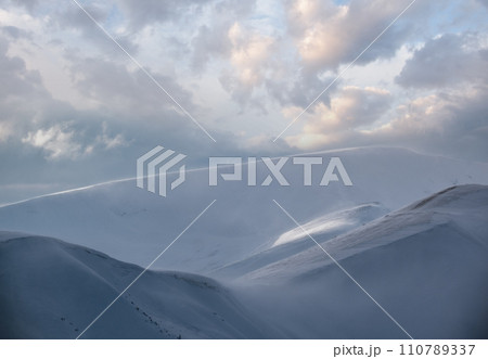 Snow covered winter mountains  in last evening sunlight. Magnificent windy dusk on tops above picturesque alpine ski resort 110789337