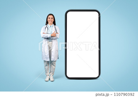 Young female doctor in white coat standing beside huge cellphone screen, pointing at blank screen, medical info display Young female doctor in white coat standing beside huge cellphone screen, pointing at blank screen, medical info display 110790392