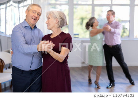 Happy smiling elderly woman enjoying impassioned merengue with male partner in latin dance salon. Social dancing concept Happy smiling elderly woman enjoying impassioned merengue with male partner in latin dance salon. Social dancing concept 110794302