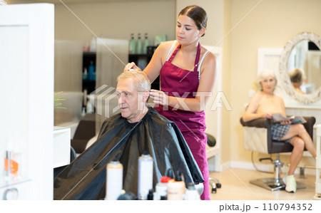 Young woman hairdresser performs haircut for elderly man using hair clipper Young woman hairdresser performs haircut for elderly man using hair clipper 110794352