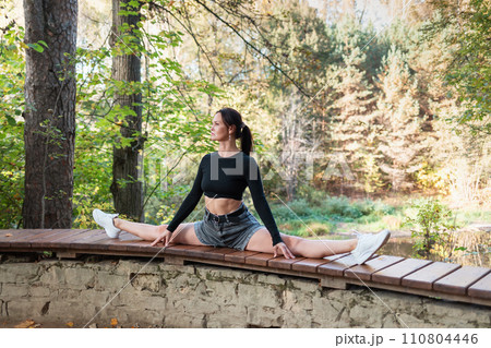 A slender woman performs the exercise Samakonasana, cross split, training in casual clothes on a sunny evening in the park, sitting on the railing of the observation deck 110804446