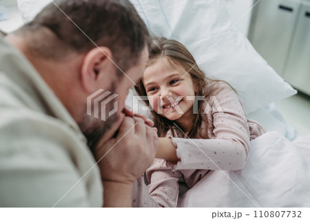 Father sitting beside daughter lying on hospital bed, playing with plush toy. Gift from dad to sick little girl in hospital. 110807732