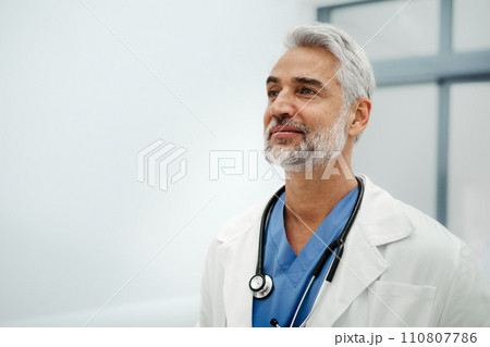 Portrait of confident mature doctor standing in Hospital corridor. Handsome doctor with gray hair wearing white coat, stethoscope around neck standing in modern private clinic, looking at camera. 110807786