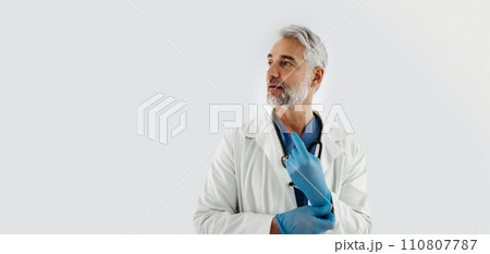 Confident mature doctor putting on medical gloves. Handsome doctor with gray hair wearing white coat, stethoscope around neck. Banner, white background with copy space. 110807787