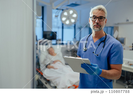 Portrait of handsome male doctor, patient in hospital bed behind. ER doctor examining senior patient, reading her medical test, lab results in clipboard. 110807788