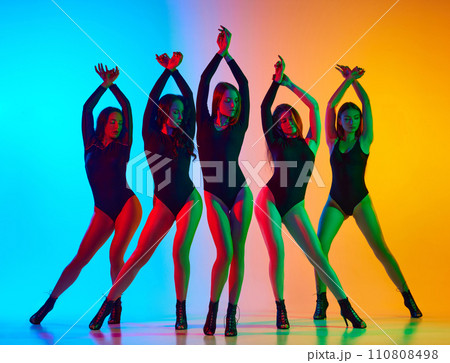 Artistic, expressive young women in black bodysuits dancing on high heels against gradient blue yellow background in neon light 110808498