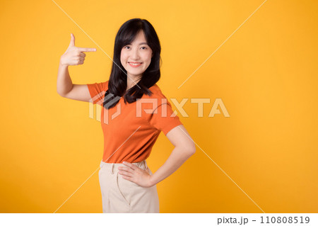 Cheerful young Asian woman in her 30s, wearing an orange shirt, points to free copy space with a happy face on yellow background. product recommending concept. 110808519