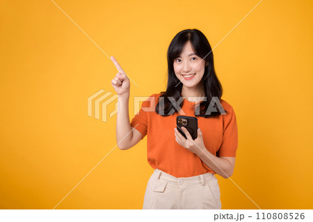 Radiate happiness with a young Asian woman in her 30s, dressed in an orange shirt, pointing to free copy space while using smartphone on yellow background. new app concept. Radiate happiness with a young Asian woman in her 30s, dressed in an orange shirt, pointing to free copy space while using smartphone on yellow background. new app concept. 110808526