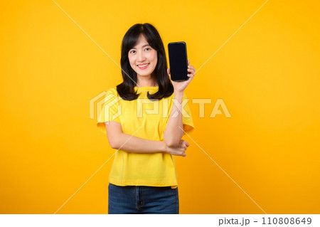 Experience joy with cheerful portrait, young Asian woman wearing yellow t-shirt and denim jeans showcases happy smile while showing smartphone screen. concept of recommended applications. 110808649