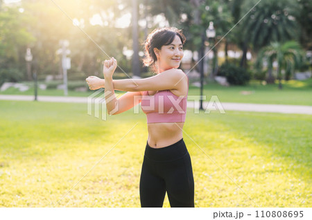 wellness and healthy lifestyle portrait 30s Asian woman in pink sportswear. preparing and stretching body and arm muscles before sunset run in the park. fitness outside and balanced life concept. 110808695