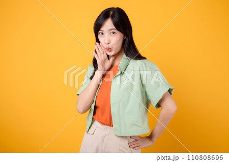 Capture young Asian woman 30s wearing a green shirt on an orange background, enthusiastically shouting with excitement. Explore the concept of discount shopping promotion with this vibrant image. 110808696