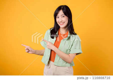 young Asian woman in her 30s wearing a green shirt on an orange shirt, pointing fingers to free copy space with enthusiasm. Explore the concept of discount shopping promotion with vibrant image. 110808710
