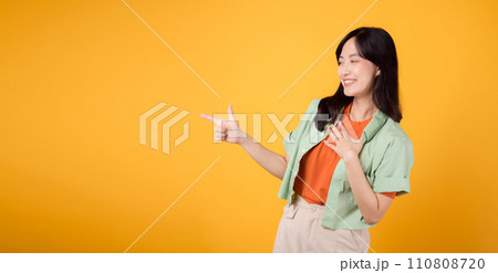 young Asian woman in her 30s, wearing a green shirt on an orange shirt, pointing fingers to free copy space. Explore the world of discount shopping promotions with this vibrant image. young Asian woman in her 30s, wearing a green shirt on an orange shirt, pointing fingers to free copy space. Explore the world of discount shopping promotions with this vibrant image. 110808720