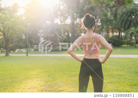 Embrace wellness living in nature as a happy, fit Asian woman in her 30s warms up her body in a public park before a sunset run, wearing pink sportswear. 110808851
