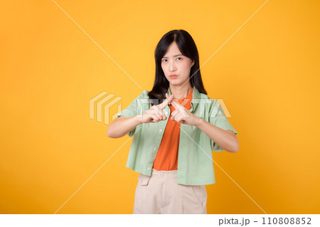 denial with a young Asian woman 30s, elegantly clad in orange shirt and green jumper. Her cross hand gesture, isolated on vibrant yellow background, represents concept of refusal and disagreement. denial with a young Asian woman 30s, elegantly clad in orange shirt and green jumper. Her cross hand gesture, isolated on vibrant yellow background, represents concept of refusal and disagreement. 110808852