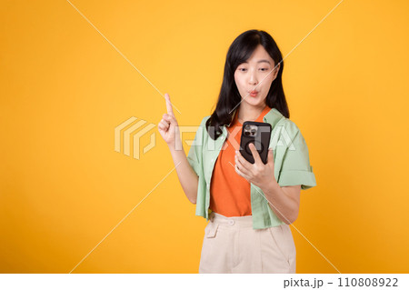 mobile apps with cheerful young Asian woman 30s, wearing orange shirt happily displays smartphone, pointing with her finger hand gesture isolated on yellow studio background. app smartphone concept. mobile apps with cheerful young Asian woman 30s, wearing orange shirt happily displays smartphone, pointing with her finger hand gesture isolated on yellow studio background. app smartphone concept. 110808922
