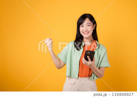 Excited young Asian woman in her 30s, wearing orange shirt and green jumper, using smartphone with fist up gesture on yellow studio background. Great news from mobile concept. Excited young Asian woman in her 30s, wearing orange shirt and green jumper, using smartphone with fist up gesture on yellow studio background. Great news from mobile concept. 110808927