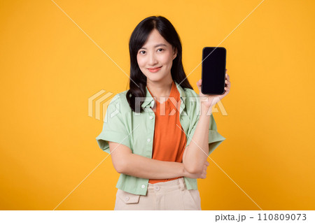 Happy young Asian woman 30s, wearing orange shirt and green jumper, displays smartphone screen on yellow studio background. New mobile application, online shopping, and new mobile device concept. 110809073