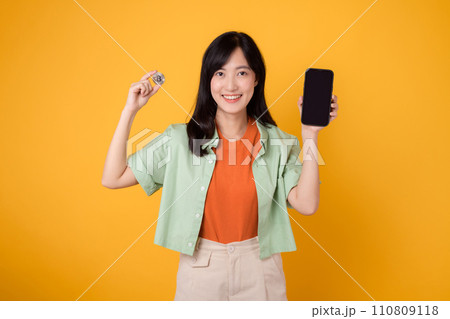 the future of finance with a captivating young Asian woman in her 30s, elegantly dressed in orange shirt and green jumper, using smartphone screen and crypto currency coin on yellow background. 110809118