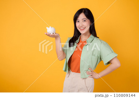 young Asian woman in her 30s, dressed in orange shirt and green jumper, showcasing piggy bank while striking an akimbo gesture on yellow background. Financial money concept. 110809144