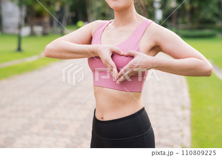 Woman jogger. young asian female happy smile wearing pink sportswear showing heart hands gesture on chest before running in public park. Healthcare wellness concept. 110809225