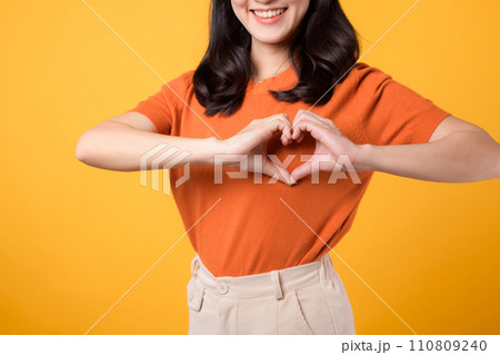 healthcare, wellness, and well-being with young Asian woman 30s wearing an orange shirt. Witness heart hand gesture on her chest against a vibrant yellow background, concept of holistic well-being. 110809240