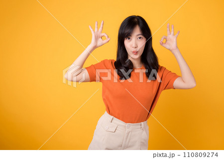 In a vibrant yellow backdrop, a young Asian woman in her 30s, donning an orange shirt, displays the okay sign gesture. Hands gesture concept. 110809274