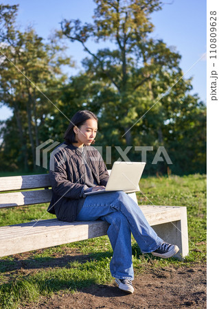 Portrait of young student, girl using laptop, sitting in park on bench, typing on computer 110809628