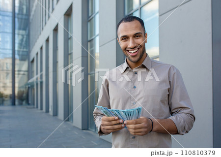 Confident African American male professional in casual wear holding money, expressing satisfaction and success outside an office building. 110810379