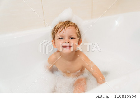 Cute little boy with foam hat taking a bath in the tub and looking at camera 110810804