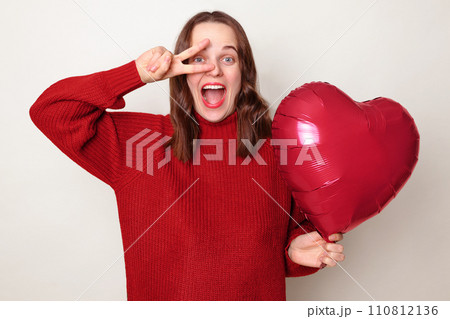 Excited festive woman with brown hair wearing red jumper making victory sign in front her eyes having fun standing isolated over gray background with heart shaped balloon 110812136