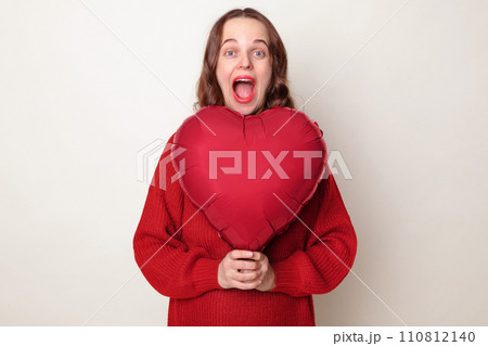 Surprised amazed Caucasian woman wearing red sweater holding heart shaped balloon isolated over gray background screaming wow loud with excited face celebrating festive event 110812140
