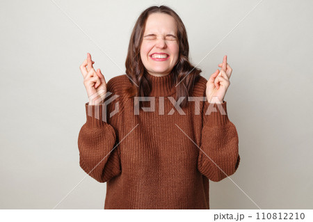 Attractive brown haired adult woman wearing brown sweater posing isolated over gray background standing with crossed fingers and closed eyes praying for good luck making wish Attractive brown haired adult woman wearing brown sweater posing isolated over gray background standing with crossed fingers and closed eyes praying for good luck making wish 110812210