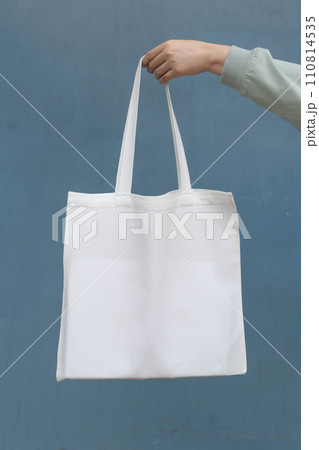 Blank white tote bag canvas fabric with handle mock up design. Close up of woman hand holding eco or reusable shopping bag on blue metal wall. No plastic bag and ecology concept. Vertical. Blank white tote bag canvas fabric with handle mock up design. Close up of woman hand holding eco or reusable shopping bag on blue metal wall. No plastic bag and ecology concept. Vertical. 110814535