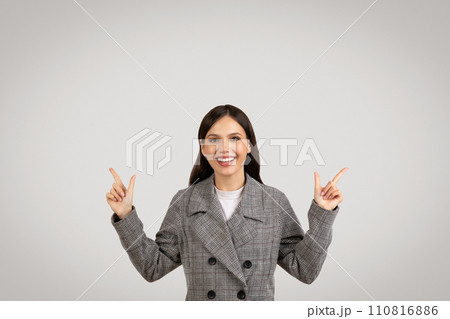 Cheerful woman in plaid jacket pointing upwards with both hands 110816886