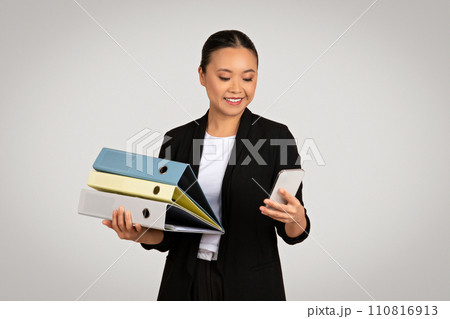 Organized Asian businesswoman carrying colorful folders and using a smartphone 110816913