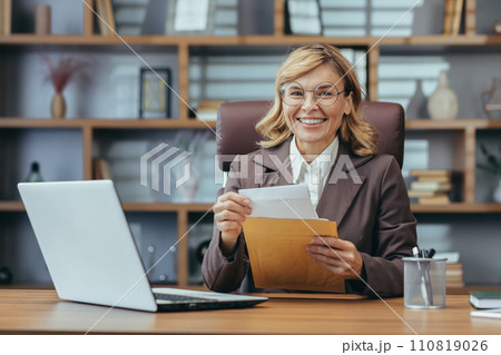 Smiling mature businesswoman with glasses working on laptop and handling documents in a well-appointed home office setting. 110819026