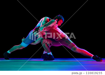 Two young athlete man, skilled wrestlers in red and blue uniform wrestling in motion against black background in mixed neon lights. Two young athlete man, skilled wrestlers in red and blue uniform wrestling in motion against black background in mixed neon lights. 110819235