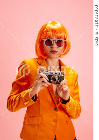 Vibrant fashion. Young woman in trendy sunglasses dressed orange blazer, with wig looking at camera and holds photo camera against pastel pink background. Vibrant fashion. Young woman in trendy sunglasses dressed orange blazer, with wig looking at camera and holds photo camera against pastel pink background. 110819303