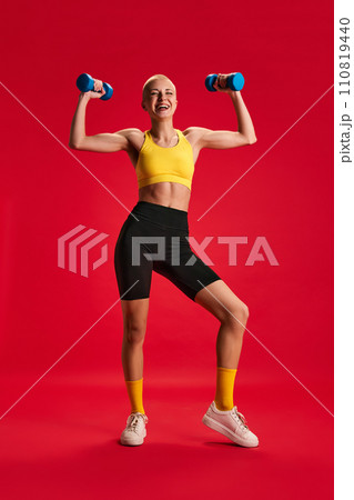 Full length portrait of woman wearing sportwear doing warming up exercises with dumbbells for arm muscles against red background. Full length portrait of woman wearing sportwear doing warming up exercises with dumbbells for arm muscles against red background. 110819440