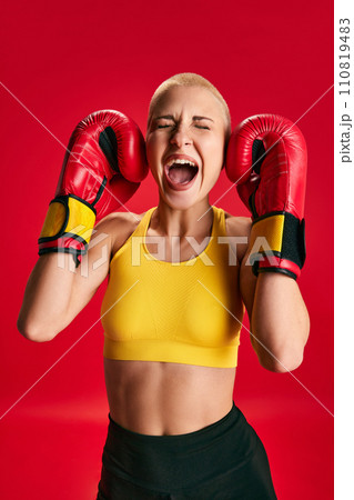 Young sporty emotional woman in sport wear and boxing gloves shouting loudly and closed eyes and ears of her self noise against red studio background. Young sporty emotional woman in sport wear and boxing gloves shouting loudly and closed eyes and ears of her self noise against red studio background. 110819483