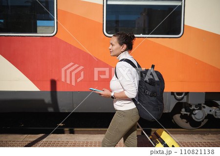 Female tourist with suitcase and backpack, walking along railway platform with smartphone, harrying up to board a train Female tourist with suitcase and backpack, walking along railway platform with smartphone, harrying up to board a train 110821138