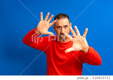 Hispanic man with beard in his 40s with his hands open in front of his face in protection mode isolated on blue studio background. Hispanic man with beard in his 40s with his hands open in front of his face in protection mode isolated on blue studio background. 110823711