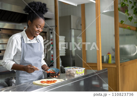 Smiling dark-skinned woman preparing pizza and looking contented Smiling dark-skinned woman preparing pizza and looking contented 110823736