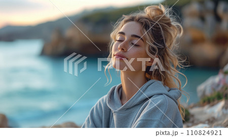 An adult beautiful girl with closed eyes in a tracksuit sits on the shore of the sea, a relaxed and happy smile on her face, close-up and copy space for the concept of health restoration An adult beautiful girl with closed eyes in a tracksuit sits on the shore of the sea, a relaxed and happy smile on her face, close-up and copy space for the concept of health restoration 110824921
