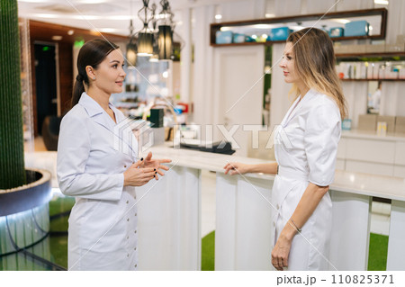 Medium shot of professional female doctor and young nurse talking, standing in clinic lobby. Physicians colleagues working together, discussing patient diagnosis in hospital corridor. 110825371