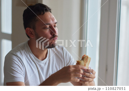 Closeup portrait of hungry man eating delicious sandwich at home on background of kitchen. Handsome male enjoying vegetarian dinner indoors. Caucasian guy eating toast in morning, slow motion. 110827051