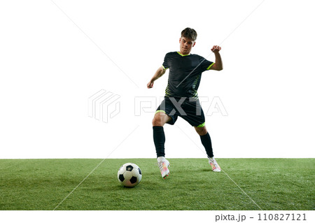 Young man in black uniform, football player playing, dribbling ball isolated over white background with grass flooring 110827121