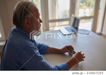 Portrait of bearded senior pensioner male with wrinkled hands counting Russian ruble banknotes sitting at home table. 110827203